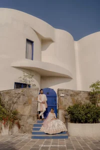 Bride in blush pink embroidered lehenga seated on blue-painted steps and groom in rose pink sherwani standing in a blue arched doorway of a white curved architectural boutique wedding venue in Rajasthan under a clear blue sky, capturing a striking architectural couple portrait