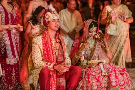 Groom in cream and gold sherwani with a deep red silk dupatta and a red and cream pearl-draped turban seated beside bride in a heavily embroidered red bridal lehenga with full gold jewellery both smiling during their wedding ceremony ritual moments at a luxury destination wedding in Udaipur Rajasthan