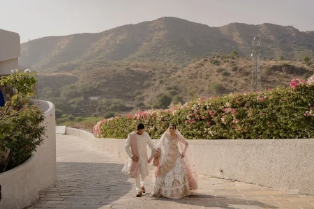 Bride in white floral embroidered lehenga with pink dupatta and groom in blush pink sherwani walking hand in hand along a stone pathway with Aravalli hills and blooming pink bougainvillea in the background at a luxury hillside destination wedding venue in Rajasthan