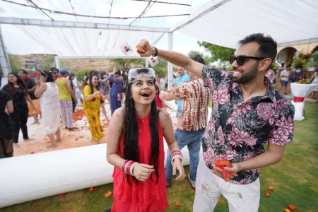 Young bride in a wet red casual kurta wearing clear protective goggles laughing with mouth open as a man in a printed pink floral shirt raises a crushed tomato about to throw it at her during a La Tomatina inspired tomato fight pre-wedding activity with multiple guests visible playing in the background at a destination wedding celebration in Rajasthan