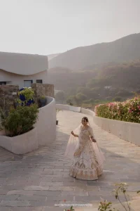 Bride in white floral embroidered lehenga with pink dupatta walking joyfully along a curved stone pathway at a luxury hillside wedding venue in Rajasthan, with Aravalli mountains and blooming bougainvillea visible in warm golden morning light