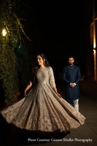 Bride in a full-length champagne beige heavily embroidered anarkali gown densely covered in small mirror work and dark floral embroidery motifs spinning with the skirt flaring out wide and her head turned away with a joyful expression against a dark outdoor night setting with lush trailing green ivy climbing a warm amber lit wall lamp on the left while the groom stands in the soft background in a dark navy blue sherwani with his hands clasped watching her at a luxury destination wedding in Rajasthan