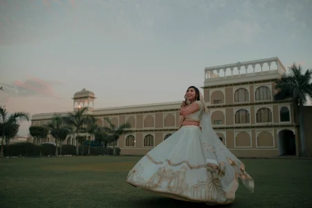Bride with a wide joyful smile spinning in a full flared white lehenga densely embroidered along the entire hem border with detailed gold zardozi depictions of Indian heritage monuments and palace architecture with a matching white and gold embroidered dupatta flowing outward and a silver diamond choker necklace at her throat photographed on a wide grass lawn at golden hour with a large multi-storey sandy beige heritage palace hotel with arched window facades white balustraded rooftop terrace and white chhatri pavilion tower glowing in warm sunset light with tall palm trees on both sides against a soft pink and pale blue dusk sky at a luxury destination wedding in Rajasthan