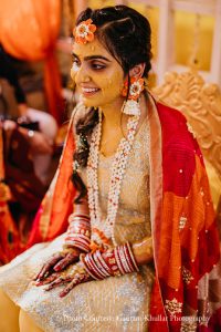 Close portrait of bride seated during haldi ceremony with bright yellow turmeric paste spread across her cheeks nose and forehead smiling softly to one side wearing a large orange fabric flower hair accessory pinned above her right ear layered white jasmine gajra garland and matching white floral earrings with mehndi-covered hands and red and white chooda bangles resting on her ivory embroidered lehenga with a warm orange silk dupatta draped over her shoulder and soft golden ambient light from behind at a luxury destination wedding haldi ceremony in Rajasthan