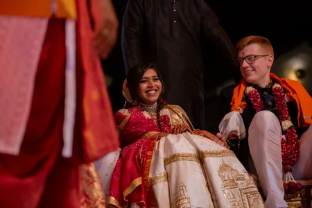 Bride in a deep magenta pink bandhani saree with a wide gold zari border and red sindoor on her forehead wearing a wide silver oxidized choker necklace and a red and white fresh flower jaimala garland beaming with a wide open smile while seated beside a red-haired groom in a dark navy sherwani with black-rimmed glasses a red tilak mark on his forehead and an orange ceremonial dupatta draped over his shoulders along with his own red and white jaimala garland with a white lehenga embroidered with gold palace and monument motifs laid across their laps during post-ceremony rituals at a luxury destination wedding in Rajasthan