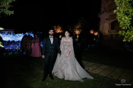 Bride in a silver and blush heavily embroidered feathered cape ball gown lehenga and groom in a dark pinstripe suit walking hand in hand across a green garden lawn at a heritage palace with guests in the background and a large glowing white floral installation under the open night sky at a destination wedding in Rajasthan