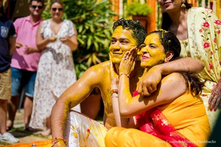 Groom shirtless in a white dhoti completely covered in bright yellow turmeric haldi paste from his face and hair to his upper body sitting beside the bride who wears an orange and yellow outfit with haldi across her face as she leans her head against his and gently cups his haldi-covered cheek with her hand both smiling contentedly while an older woman's hands are visible at the top of the frame and a mix of Indian and international Western guests including a man in a pink shirt and a woman in a floral dress stand in the softly blurred lush green garden background at a luxury destination wedding haldi ceremony in Rajasthan