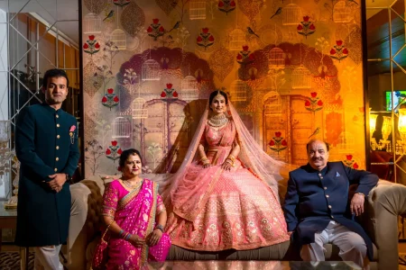 Bride in a heavily embroidered blush pink bridal lehenga with a large flared skirt gold maang tikka layered polki necklace and flowing pink net dupatta seated regally in the center as her brother in a dark teal embroidered bandhgala stands to the left and her mother in a bright pink silk saree and father in a dark navy suit sit on either side of her in front of a richly lit gold and maroon ornate Mughal birdcage and bird illustrated mural backdrop at a luxury destination wedding in Rajasthan