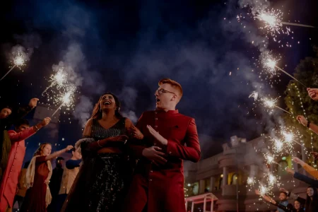 Close-up dramatic photograph of bride in a dark navy black heavily sequined lehenga with sheer embroidered short sleeves laughing loudly with eyes wide open while a red-haired groom in a bold deep crimson red sherwani with silver buttons and black-rimmed glasses looks up in wide-eyed excitement as sparklers held by guests on both sides cross directly above them sending cascading white sparks through the air with thick blue and white smoke dramatically billowing upward into the dark night sky and a softly illuminated heritage palace structure visible in the background at a luxury destination wedding sangeet in Rajasthan