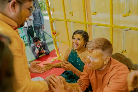 Bride in a teal dark green halter top completely covered in thick bright yellow turmeric haldi paste on her face neck and arms laughing openly while holding out her haldi-smeared hands toward someone off-frame beside a red-haired groom in an orange kurta whose face is turned away and also covered in haldi paste seated together on the red-carpeted floor of a yellow canopy-covered ceremony space decorated with yellow fabric walls and marigold tassels with a wedding photographer crouching in the background capturing the moment at a luxury destination wedding haldi ceremony in Rajasthan