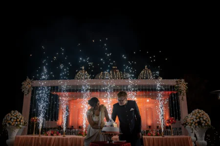 Couple bending together over a white fondant wedding cake decorated with a small cake topper on a table draped in pale blush linen with the bride in a gold champagne embroidered saree leaning in and the red-haired groom in a dark navy sherwani with glasses standing beside her as a dramatic silver cascading sparkler waterfall curtain falls behind them and starburst cold pyrotechnics shoot upward above a large decorative lattice white stage backdrop illuminated in deep warm orange with tall white urn floral arrangements on both sides of the cake table against a completely dark night sky at a luxury destination wedding reception in Rajasthan