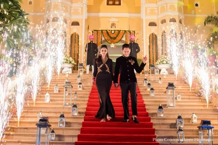 Couple walking hand in hand down a wide red-carpeted palace staircase lined on both sides with tall silver lanterns while silver cold pyrotechnic spark fountains shoot dramatically upward around them with the woman in a black floor-length gown with gold star-motif embroidered detailing and red chooda bangles and the man in a black bandhgala suit with a large ornate gold brooch pointing one finger upward while two attendants in deep pink turbans and dark sherwanis stand at the top of the stairs against a backdrop of orange marigold garland swags and the illuminated warm golden palace facade at a luxury destination wedding reception in Rajasthan
