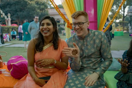 Bride in a peach coral raw silk saree with a wide gold border and both forearms completely covered in dense dark reddish-brown mehndi laughing broadly beside a red-haired groom wearing black-rimmed glasses and a blue-grey printed kurta with small gold buta motifs who holds up his palm to show a freshly applied sun motif henna design while seated outdoors on colorful cushions under a vibrant canopy of multi-colored drapes in green hot pink and orange with thick orange marigold garland swags and lush garden trees and a pale pink heritage building visible in the blurred background at a luxury destination wedding mehendi ceremony in Rajasthan