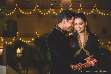 Couple in matching black and gold embroidered outfits photographed outdoors at night with the man in a black sherwani featuring gold shoulder and collar embroidery and jeweled buttons leaning his forehead gently toward the woman who wears a black outfit with dramatic gold embroidered lapels red and pink chooda bangles mehndi-covered hands dangling gold jhumka earrings and vivid blue eye makeup with rows of warm golden string light bokeh glowing softly across the entire background and a pale heritage palace facade barely visible in the distance at a luxury destination wedding reception in Rajasthan