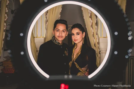 Couple dressed in matching all-black outfits with intricate gold embroidery photographed framed within a glowing circular LED ring light with the man on the left in a black bandhgala with gold collar trim and jeweled buttons and the woman on the right in a black outfit with bold gold embroidered lapels both looking directly into the camera with ivory cream curtains with gold trim and an ornate carved column softly visible in the background inside a heritage palace interior at a luxury destination wedding in Rajasthan