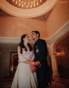 Bride in a blush pink heavily embroidered lehenga with intricate orange mehndi gloves on both arms laughing up at groom in a dark navy embroidered bandhgala suit as they hold each other close inside a grand heritage palace interior with a large gold coffered ceiling chandelier and delicate floral patterned warm amber walls with white arched columns visible in the background at a luxury destination wedding in Rajasthan