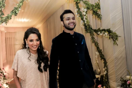 Woman in champagne beige sequin top with open wavy hair and statement diamond necklace and man in dark navy blue bandhgala smiling together under a leafy green floral arch at an elegantly decorated indoor wedding reception at a destination wedding venue in India