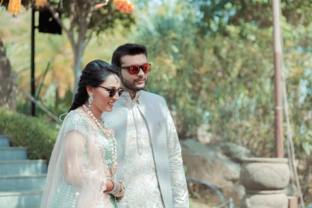 Bride in aqua mint sequin lehenga with layered pearl jewelry and groom in silver embroidered sherwani both wearing stylish sunglasses walking together at an outdoor pre-wedding event with lush green garden backdrop at an inter-community destination wedding in India