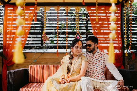 Bride in an ivory cream embroidered lehenga with a side-parted braid adorned with a large orange flower hair accessory and layered white floral gajra garland necklace with mehndi-covered hands resting in her lap gazing downward with a serene expression while the groom beside her in a white kurta printed with small red elephant motifs and dark sunglasses drapes his arm around her as they sit together on a red and white candy-striped cushioned sofa seat under a vibrant orange fabric canopy decorated with hanging orange and yellow bead tassel strings diamond-shaped colorful pendants and a black and white horizontal slatted backdrop with green tropical vines above at a luxury destination wedding mehendi ceremony in Rajasthan