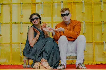 Bride in a slate grey embroidered salwar suit with dark round sunglasses and both hands densely covered in dark reddish-brown mehndi holding out her palm to display an intricate circular sun motif henna design while a red-haired groom beside her in a burnt orange kurta with white trousers and dark rectangular sunglasses points at the design smiling as they sit on carved wooden chairs in front of a bright yellow fabric grid backdrop with small marigold tassels hanging from each row at a luxury destination wedding mehendi ceremony in Rajasthan