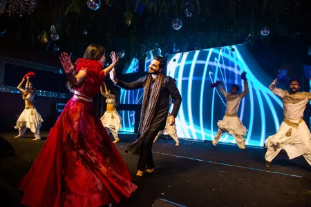 Bride in a red floral printed full-length gown with large sculptural red ruffle shoulder and mehndi hands reaching out toward groom in a black embroidered sherwani with a long grey draped stole as he leans in smiling broadly during a choreographed couple sangeet stage performance with four backup dancers in white dhoti style outfits visible on either side and a vivid blue swirling digital LED screen backdrop with mirror ball lights overhead at a luxury wedding sangeet event in India