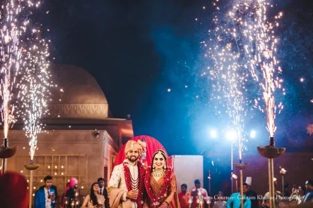 Wide nighttime photograph of newlywed couple standing together smiling at the camera with the groom in a cream ivory sherwani with a peach safa turban and layered red and maroon jaimala garland beside the bride in a deep red heavily embroidered bridal lehenga with a matching red dupatta and layered gold kundan bridal jewelry as tall silver fountain pyrotechnics erupt dramatically on both sides and large aerial firework bursts spread across the entire dark blue night sky above them with a sandstone heritage dome structure lit in warm amber on the left bright blue stage lights and a red draped ceremony backdrop with staff and guests softly visible at the edges at a luxury destination wedding in Rajasthan