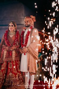 Bride in a deep crimson red heavily embroidered lehenga with intricate gold zardozi work and a matching red dupatta wearing a layered gold polki and kundan necklace set large gold jhumka earrings and a gold maang tikka standing with hands clasped and looking forward beside a bearded groom in a cream off-white embroidered sherwani with a peach safa turban a large gold sarpech brooch and a layered deep red and maroon jaimala garland also looking ahead as cascading silver cold pyrotechnic waterfall sparks fall in a dense curtain on the right side of the frame against a completely dark night background at a luxury destination wedding in Rajasthan