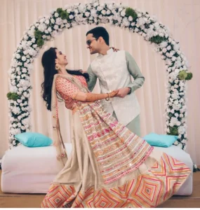 Bride in a multi-color phulkari mirror work lehenga with ivory and orange geometric border and gold dupatta twirling with arms around groom in a mint green kurta and white embroidered jacket with sunglasses both gazing at each other with smiling expressions in front of a lush circular white carnation and green flower arch with white daisy sprays and a cream draped fabric backdrop with teal cushioned white bench seating at a daytime outdoor wedding event in Rajasthan