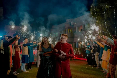 Bride in a dark navy black heavily sequined lehenga with sheer embroidered sleeves laughing with mouth wide open as she walks arm in arm with a red-haired groom in a bold deep red sherwani with gold buttons and black-rimmed glasses who looks ahead in mock surprise while two parallel lines of guests on either side hold up lit sparklers creating a tunnel of white sparks arcing overhead with blue-toned smoke drifting upward into the dark night sky and a pale heritage palace building and string lights softly visible in the background at a luxury destination wedding sangeet in Rajasthan