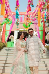 Bride in mint green sequin lehenga with blush pink dupatta and groom in silver embroidered sherwani with red tinted sunglasses walking hand in hand under a vibrant multicolored tassel and pompom canopy at their outdoor mehendi ceremony at a destination wedding in India