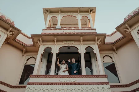 Wide low-angle upward shot of a couple standing on the ornate carved scalloped arch balcony of a grand pink terracotta and cream heritage palace with the bride in a white saree and silver choker necklace raising her mehndi-covered hand in a wave while the red-haired groom in a dark navy bandhgala sherwani with glasses beside her also waves outward with the palace facade featuring multiple layers of decorative carved arches scalloped detailing corbelled brackets and terracotta-toned borders filling the frame dramatically against a pale blue sky at a luxury destination wedding in Rajasthan