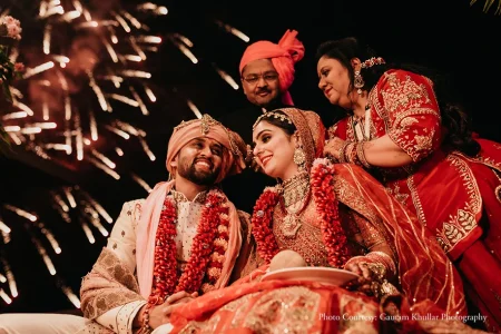 Low-angle close-up photograph of groom in a cream ivory embroidered sherwani with a peach safa turban and layered red flower jaimala garland laughing broadly while looking at the bride beside him who wears a deep red heavily embroidered bridal lehenga with a layered gold kundan necklace and gold maang tikka smiling at him as a woman in a deep red and gold embroidered saree stands directly behind them leaning in warmly and a man in a coral pink silk sherwani with matching turban stands further behind while cascading silver pyrotechnic sparks burst dramatically against the dark night sky above at a luxury destination wedding ceremony in Rajasthan