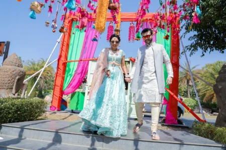 Bride in aqua blue sequin mirror work lehenga with sunglasses and groom in white embroidered sherwani with red tinted sunglasses walking hand in hand under a vibrant multicolored fabric and tassel mandap gate at their outdoor mehendi ceremony at an inter-community destination wedding in India