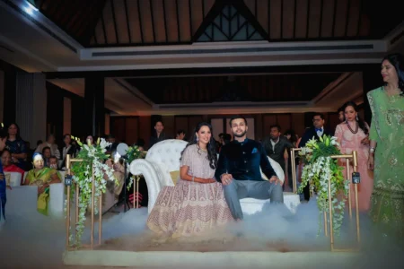 Couple seated on a white tufted throne chair surrounded by dramatic dry ice fog effect at their wedding reception, woman in mauve embroidered sequin lehenga and man in dark navy bandhgala with grey trousers, flanked by white lily flower arrangements and a green garland arch, with guests seated in the audience at a luxury wedding reception in India