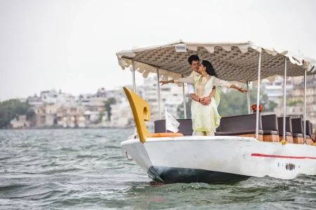 Couple sharing a playful and romantic moment standing on a traditional covered white wooden boat sailing on Lake Pichola with the Udaipur city ghats and buildings visible in the background, bride in a pastel mint green embroidered suit with mehndi designs on her hands, during a pre-wedding photoshoot in Udaipur Rajasthan