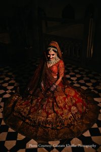 Moody high-contrast bridal portrait shot from directly above with the bride in a sweeping deep crimson red heavily embroidered lehenga with intricate gold zardozi and thread work covering the entire skirt including detailed arch motifs along the hem border seated with the skirt fanned out fully around her in a circle on a classic black and white checkered marble floor wearing a wide diamond and pearl polki choker necklace gold maang tikka large statement earrings and dark kohl-rimmed eyes with a matching red dupatta draped over her head looking downward with a composed expression in dramatic directional low light against a near-black background at a luxury destination wedding in Rajasthan