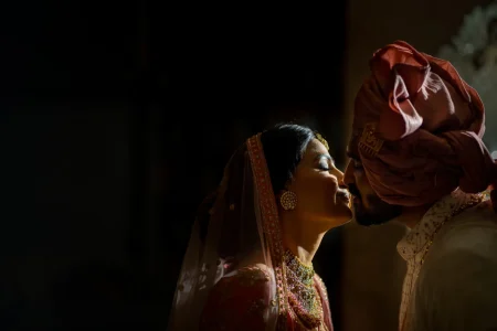 Intimate and dramatic close-up of bride in a red embroidered bridal lehenga with heavy gold polki necklace and large disc earrings closing her eyes as she lifts her face toward groom in a terracotta rust safa turban and cream sherwani who leans in close both lit by a single shaft of warm directional light against a deep black background at a luxury heritage palace wedding in Rajasthan