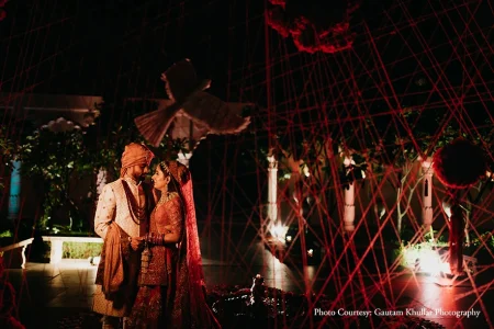 Groom in a cream off-white embroidered sherwani with a peach safa turban and deep maroon layered jaimala garland gazing at the bride beside him who wears a deep crimson red heavily embroidered lehenga with a matching red dupatta and gold bridal jewelry both standing close and looking at each other inside a dramatic artistic installation of criss-crossing diagonal red strings stretched at sharp angles filling the entire frame from floor to ceiling with suspended red floral balls and a white sculpted bird in mid-flight visible above them a dark outdoor garden setting with softly illuminated white columns lush green foliage and warm red ambient ground lighting at a luxury destination wedding in Rajasthan