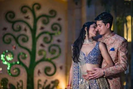 Couple sharing an intimate close portrait moment at night, woman in a royal blue and silver embroidered lehenga with diamond choker necklace and man in a peach rose gold floral brocade sherwani, with an illuminated decorative green tree scroll art installation glowing warmly in the background at a destination wedding engagement event in Udaipur