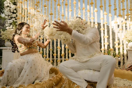 Bride and groom playfully throwing dried flower petals at each other during their haldi ceremony, bride laughing in a yellow embroidered crop top with white lehenga and groom in white kurta, with marigold string curtain backdrop at a destination wedding in Rajasthan
