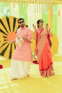 Groom in pink kurta with traditional white mundu and bride in orange peach silk saree with gold temple jewellery and sunglasses performing an expressive and comical dance together in front of a large yellow marigold sunflower-style floral backdrop at a South Indian wedding celebration in India