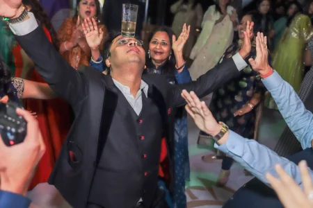 A wedding guest in a dark suit and waistcoat balancing a full glass of liquid on his forehead with arms raised wide while dancing eyes closed and head tilted back in a crowd of cheering and laughing guests at a lively and energetic wedding reception dance celebration in India