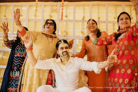 Groom in a white kurta seated at the center of an exuberant haldi ceremony with bright yellow turmeric paste streaked across his face and hair raising both arms wide open in a joyful celebratory gesture while laughing broadly as three women stand directly around him all with arms raised and laughing including one in a mustard yellow embroidered salwar kameez with dark sunglasses one in a coral orange brocade salwar and one in a deep pink and gold silk saree with a small gold maang tikka all in front of a white wooden grid frame backdrop with hanging yellow bead tassels at a luxury destination wedding haldi ceremony in Rajasthan