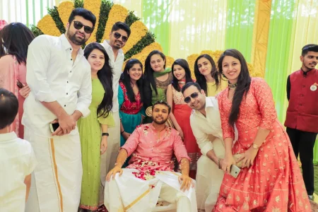 Groom in pink kurta with white mundu seated covered in rose petals and haldi paste surrounded by a large group of smiling friends and family in colorful ethnic wear posing together in front of a large marigold floral fan installation with yellow and green decor at a destination wedding in India
