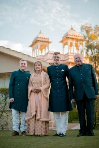 Four family members posing together on a manicured green lawn in front of a soft pink terracotta heritage palace with ornate white carved chhatri pavilion towers and a pale blue sky above with three men wearing matching deep teal dark green bandhgala sherwanis with brass gold buttons and white trousers including a groom at center in glasses and an older gentleman on the right in a matching teal western-cut blazer while an older woman in the center wears a delicate blush peach embroidered anarkali suit with a sheer embroidered dupatta and gold jhumka earrings at a luxury destination wedding in Rajasthan
