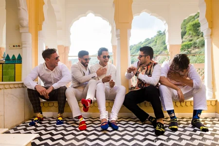 Five men in a mix of white shirts white linen suit and a colorful printed waistcoat laughing and joking candidly while seated along a low marble ledge on an open palace terrace all displaying mismatched colorful novelty superhero-themed socks prominently with Aravalli hills and dense green trees visible through white arched columns in the background a bold black and white chevron zigzag tile floor underfoot and a teal decorative tile panel to the left at a luxury destination wedding in Rajasthan