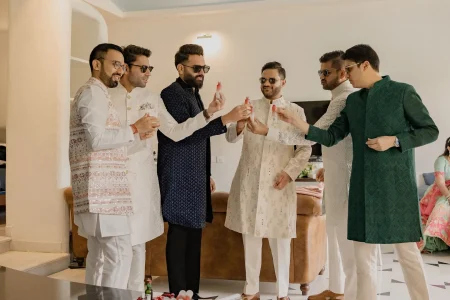 Six groomsmen wearing colorful ethnic sherwanis and sunglasses toasting with small bottles during pre-wedding celebrations inside a luxury venue room, capturing the candid fun energy of a multi-day destination wedding in Rajasthan