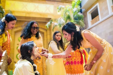 Bride seated at her haldi ceremony in a cream outfit with jasmine hair garland and gold jewellery being playfully fed by a smiling female friend leaning in closely while other women in yellow outfits watch and smile, in an outdoor courtyard at a destination wedding in Udaipur Rajasthan