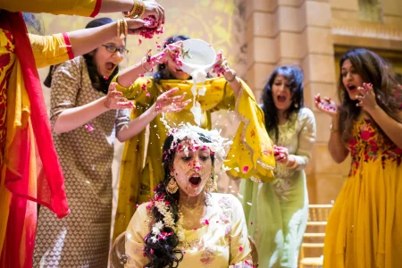 Bride seated at haldi ceremony in a cream embroidered suit reacting in shock as a friend pours a bowl of milk over her head while other women in yellow and mustard outfits laugh and throw rose petals around her, captured in a perfectly timed candid moment at a destination wedding haldi ceremony in Udaipur