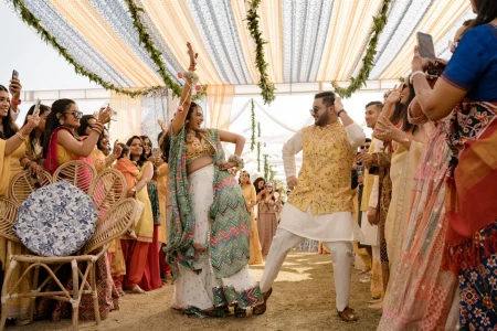 Bride in multicolored mirror work lehenga and groom in a floral yellow waistcoat over white kurta dancing energetically during their haldi ceremony entry as cheering guests form a corridor around them under a colorful canopy at a destination wedding in Rajasthan